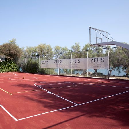 Dolce By Wyndham Athens Attica Riviera Hotel Vravrona Exterior photo The photo shows a basketball court with a reddish surface. There are two basketball hoops on either side of the court, surrounded by a fence that has banners displaying the word "ZEUS." In the background, there are trees, and the court appears to be 