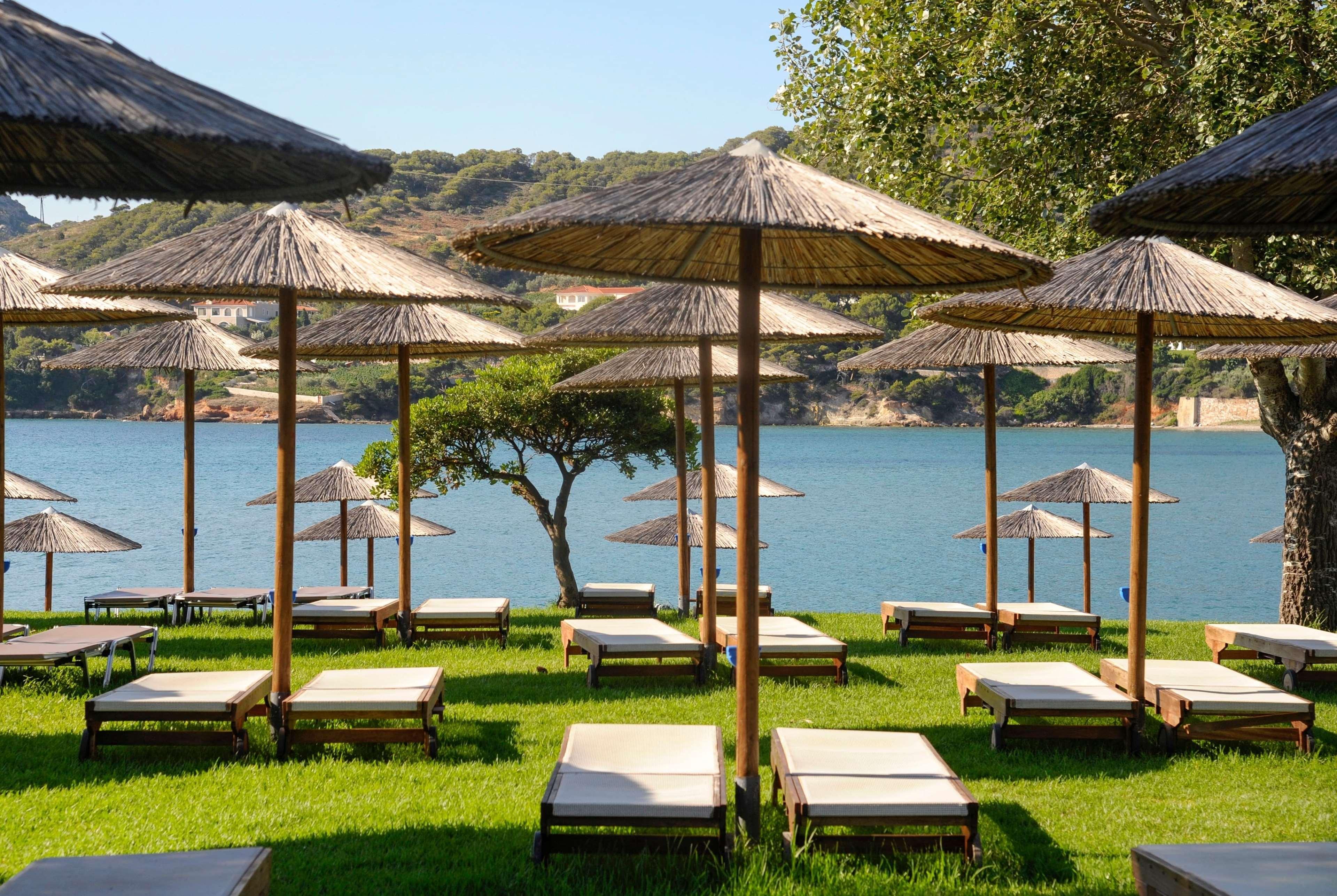 Dolce By Wyndham Athens Attica Riviera Hotel Vravrona Exterior photo The photo shows a serene beach scene with several lounge chairs arranged on a green lawn. Each lounge chair is shaded by a traditional straw umbrella, providing a relaxing spot to enjoy the view. In the background, there is a calm body of water, like