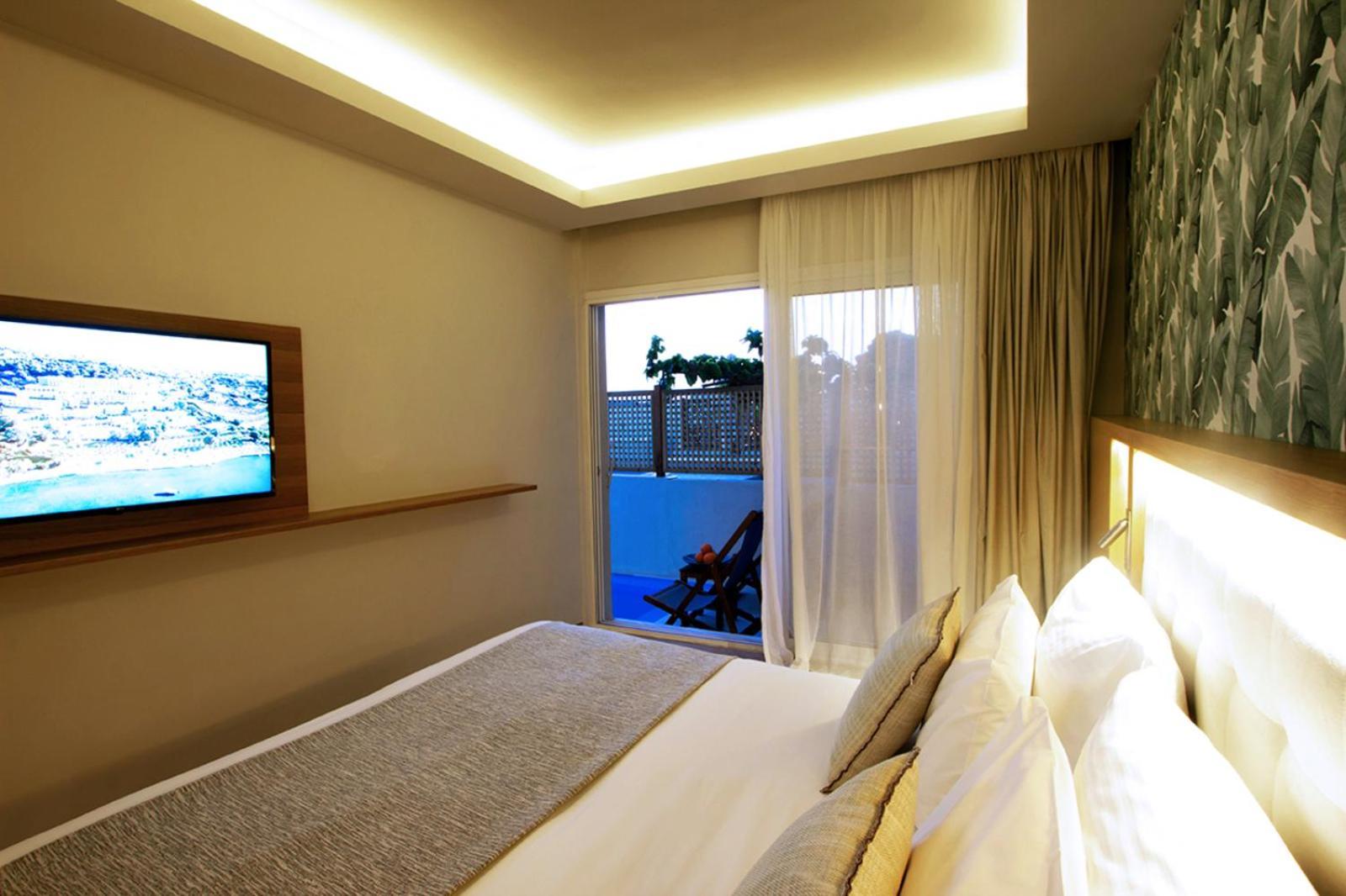 Dolce By Wyndham Athens Attica Riviera Hotel Vravrona Exterior photo The photo shows a modern hotel room featuring a large bed with white linens and decorative pillows. There's a mounted flat-screen television on the wall opposite the bed. Natural light enters the room through sheer curtains, leading to a balcony or o